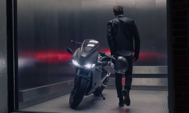 A man walks into a silver room and stands next to a Zero SR/S motorcycle with his back to the camera.