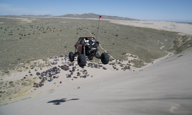 A UTV sails through the air over a steep decline on a sand dune. Many people and vehicles look like ants at the base of the hill.