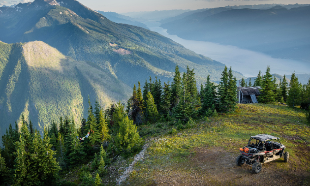 An ATV is parked on a green mountaintop. Another mountain and a river are in the background.