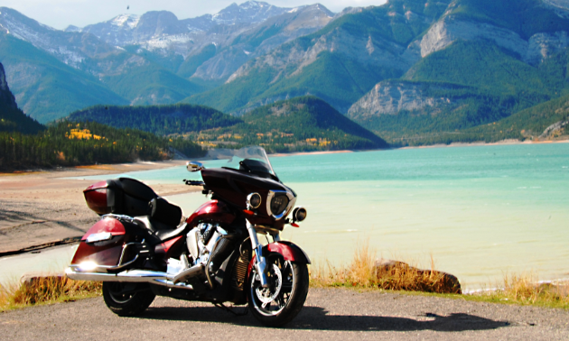A maroon motorcycle shines in the sunshine in the foreground while a turquoise lake is in the background, followed by green, jagged mountains.