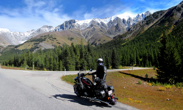Don Morberg stands next to his motorcycle at the side of the road amidst towering mountains.