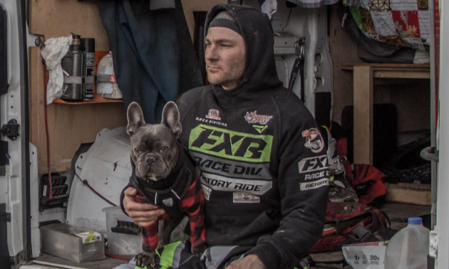 Dustin Labby sits on the end of a trailer with a dog on his lap.