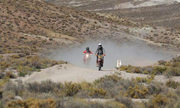 Two motorcycles drive towards the camera on a desert track.
