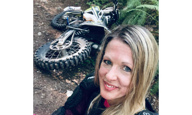 Jennifer Boulet smiles for a selfie with her dirt bike laying down behind her.