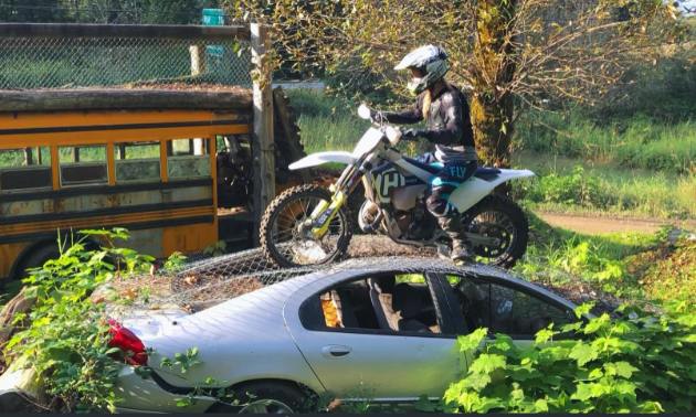 A dirt biker on top of a grey car in an enduro park. There is a school bus and tree in the background.