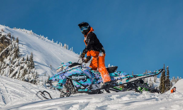 A man in orange pants stands up at the controls of a turquoise and purple snowmobile.