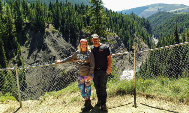Jody Stevens and her husband, John, stand in front of a fence with Elkford’s mountains in the background.