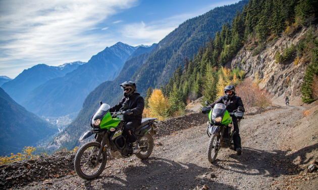 Two motorcyclists make their way up a mountain along a dirt road.
