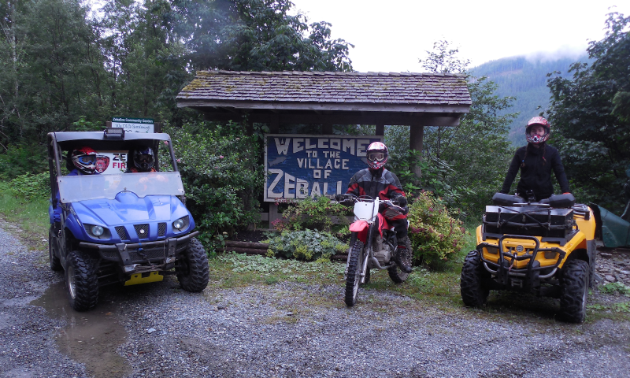 A side-by-side, dirt bike and quad line up for a photo in front of a “Welcome to Zeballos” sign. 