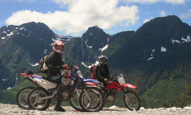 Two motorcyclists pose for a photo in front of towering mountains under a blue sky with few clouds. 