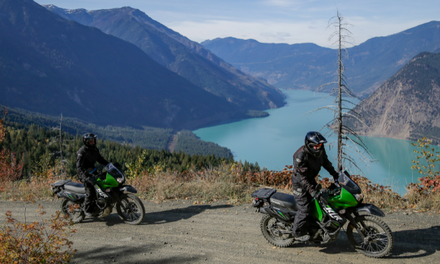 Motorcyclists ride a dirt road above Seton Lake down in a valley between mountains.
