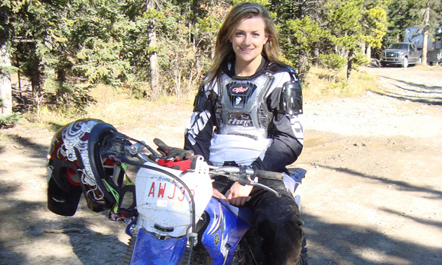 Photo of a girl with long brown hair in her mid twenties sitting on a blue dirt bike. 