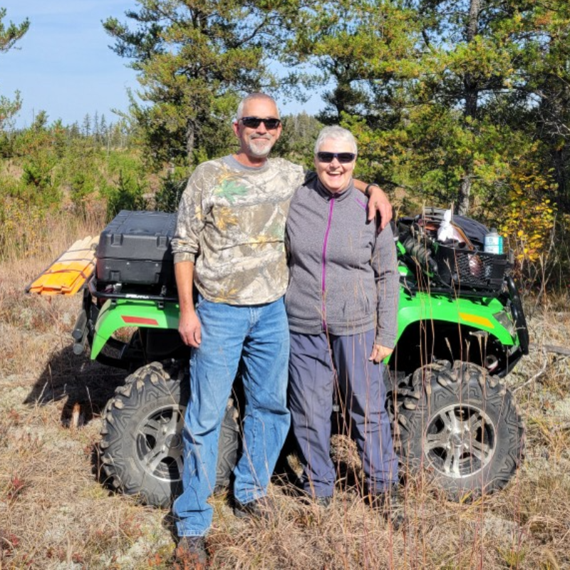 Gary Hora wears sunglasses and camo gear with his arm around Joan, who also wears shades and is sporting a bright smile. They stand in tall brown grass in front of a green ATV.