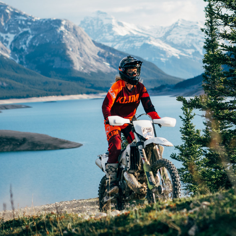 Gloria Cunningham rides a dirt bike up a mountain with a lake and more mountains in the background. 