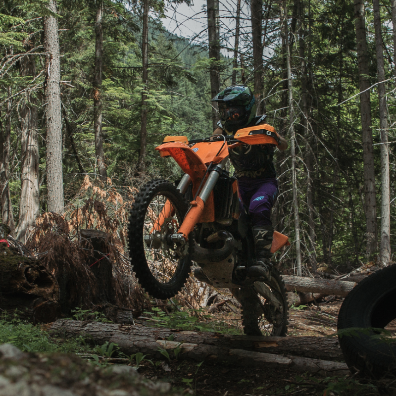 Kirsten Patton pops a wheelie while bouncing over logs in the woods on her KTM dirt bike. 