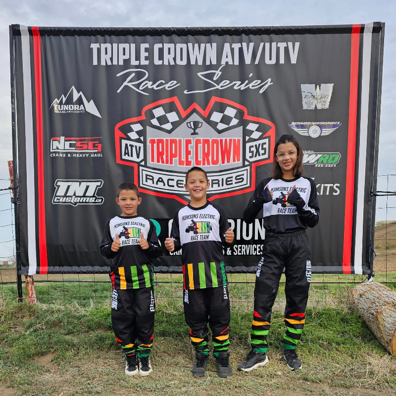 (L to R) Theo (age 8), Isaac (age 10) and Savannah (age 12) Kumson. They wear black clothes with their logo on their chests, and red, green and yellow lines wrapped around their ankles. 