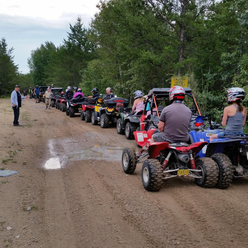 A row of ATVers line up and listen to instruction from a man standing beside the trail. 