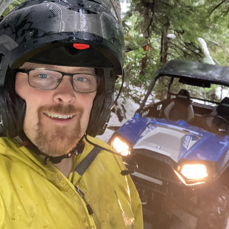 Dillon Baycroft wears a yellow jacket, black rimmed glasses and a black helmet in front of his blue side-by-side ATV with its lights on.