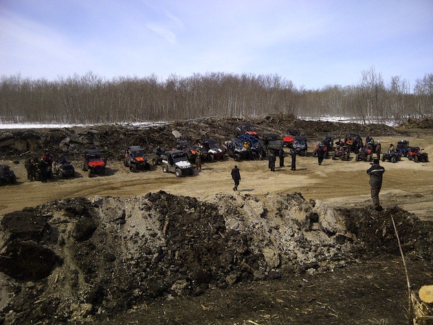 A picture of ATVers all standing around their machines waiting for the 1st ride of the season.  There are sand dunes behind them, and in the foreground. 