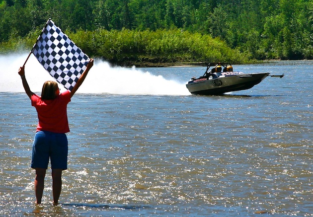 In the forground there is a women standing ankle deep in the water with a checkered flag at the finish line.  On the lake a boat is speeding past. 
