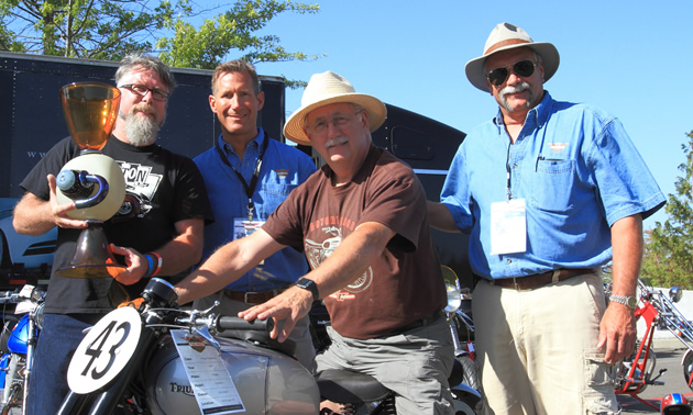 Picture of motorcycle enthusiasts, (one sitting on a motorcycle) and man holding trophy. 