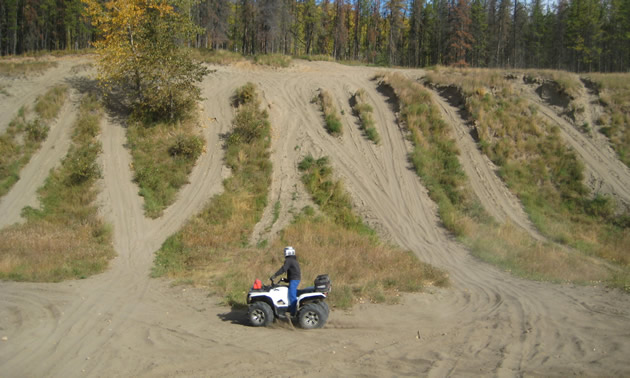 A ATV rider sits at the bottom of a dune looking up at the trails and the blue sky.