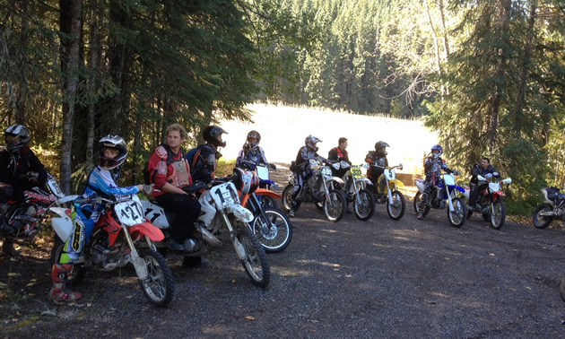 A row of riders on off-road bikes are lined up on a gravel drive in front of trees and a lake.