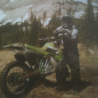 A young man stands in front of forest a mountains with a lime green dirt bike.
