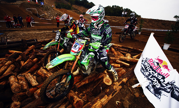 Two riders on bright green bikes try to navigate over a pile of logs.