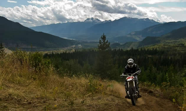 A man on a dirt bike comes around the corner with a view panorama of mountains behind him.