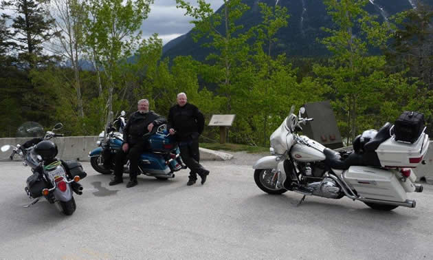 A man with white hair and beard sits on a blue road king motorcycle while another man stands next to him. Two other bikes are in the foreground.