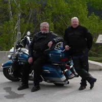 A man with white hair and beard sits on a blue road king motorcycle while another man stands next to him. Two other bikes are in the foreground.