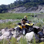 A rider on a yellow quad navigates through a field of boulders. Behind him is a rolling green hill.
