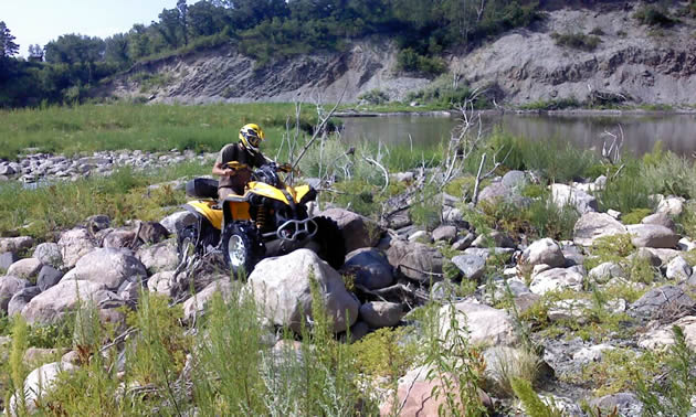 A rider on a yellow quad navigates through a field of boulders. Behind him is a rolling green hill.