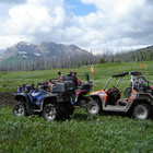 Two ATVs and a Razor are parked in an expanse of green field with a mountain off in the distance.
