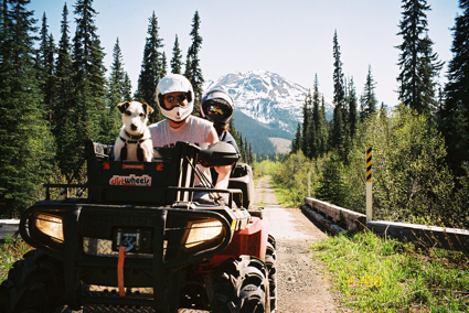 A man and his dog sit and pose for the camera on an ATV on a trail through the forest and mountains