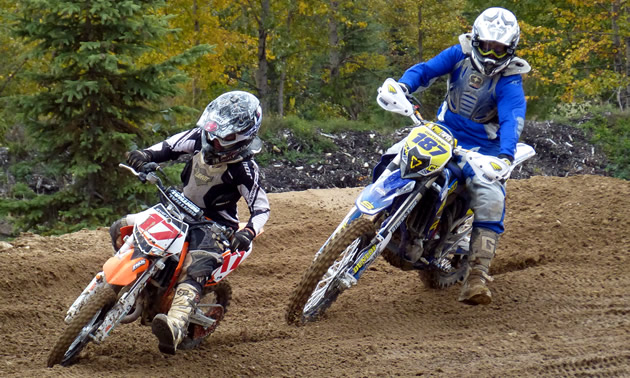 Two motorcyclists race around a dirt track in the woods.
