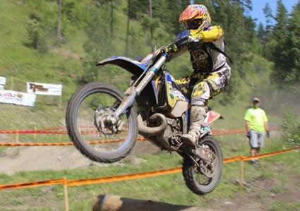 Photo of a guy jumping a dirt bike over a pile of logs on a dirt track. 