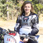 Photo of a girl with long brown hair in her mid twenties sitting on a blue dirt bike. 