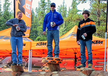 Photo of three men standing on a log podium with drinks in their hand and orange tents in the background. 