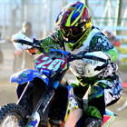 A guy in blue and green riding gear jumping a blue dirt bike over a log on a track. 