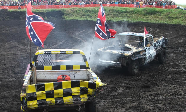 A yellow truck, decked out with confederate flags, smashes into a black truck in front of a large crowd.