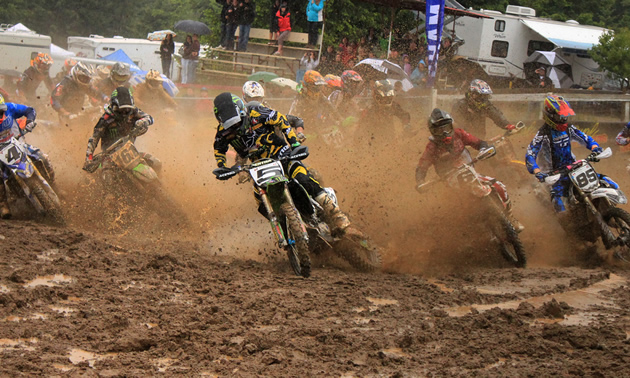 Photo of a group of guys on dirt bikes, sliding around the corner and flinging up mud on a motocross track. 