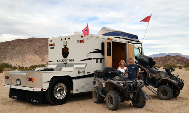 A man and woman standing by their modified toy hauler with ATVs. 