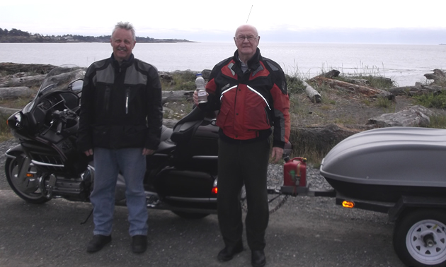 Photo of an older gentleman and a middle-aged man standing in front of a BMW motorcycle with a trailer attached to it. The rig is parked beside the ocean. 