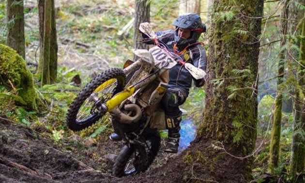 A dirt biker ascends a hill in the woods by doing a wheelie. 