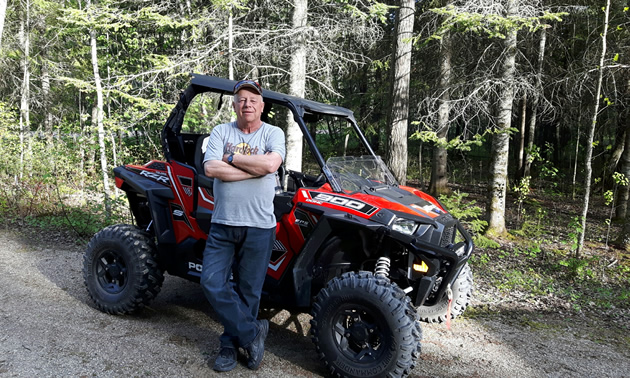 An older gentleman standing beside a red quad in the forest. 
