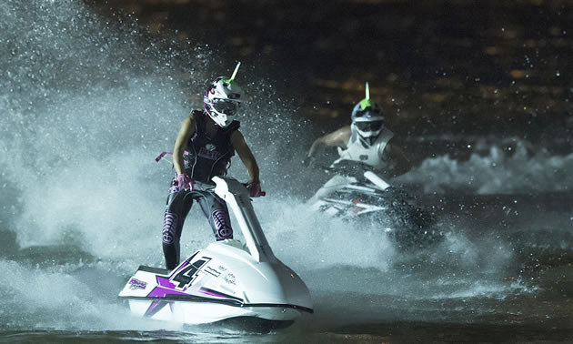 Photo of two people standing on jet skis and racing them in the dark. 
