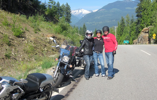 Three motorcycle riders standing by a Harley Davidson on the side of the road. 