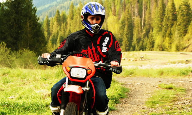A boy in a blue helmet riding wearing a black and red long sleeve shirt riding a orange dirt bike on a grassy trail in the backcountry. 